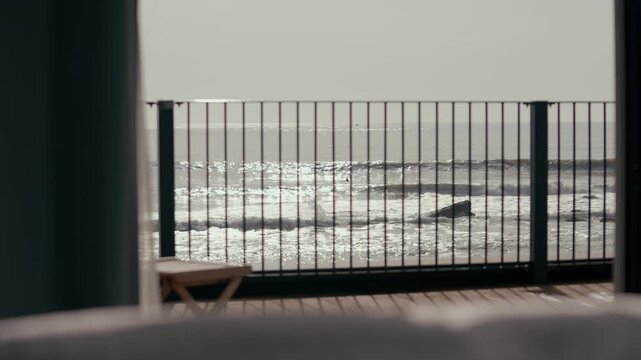 Ocean view from wooden deck with metal railing. Waves crash against shore in soft focus. Outdoorsy calm atmosphere with muted tones. Suitable for travel, vacation, or coastal relaxation themes