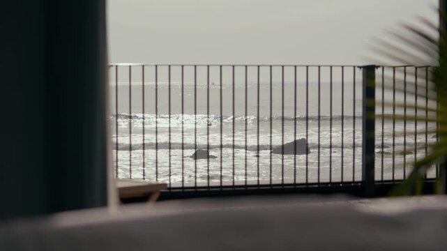 View through balcony railing shows ocean waves. Palm fronds frame right side. Sky overcast, light gray tones. Sea surface reflects soft daylight. Railing bars create graphic pattern