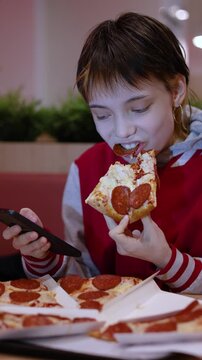 Social media takes over as a teenage girl sits in a fast food restaurant, phone glued to her hand, enjoying her pizza slice without ever looking up from the screen.