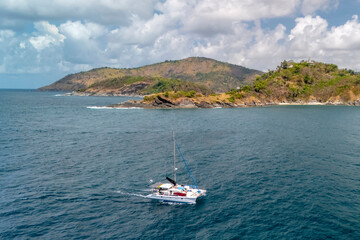 Obraz premium A sleek white catamaran rests on calm, deep blue water. A vibrant green tropical coastline with rocky areas and a small sandy beach forms the backdrop under bright daylight