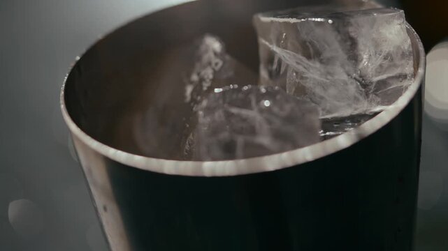 Close-up of ice cubes in a metallic shaker. Smooth, reflective surface catches light softly. Ice edges glisten with condensation. Shaker appears professional, ready for cocktail mixing