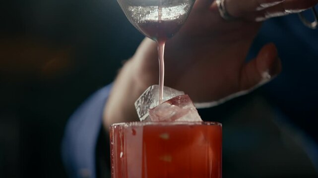Barman pours red cocktail through strainer into glass with ice. Close-up shows liquid flowing smoothly, highlighting bartending skill. Soft lighting and blurred background suggest cozy bar atmosphere