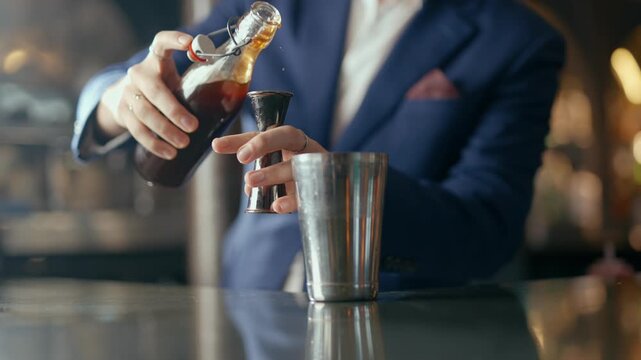 Barman pours dark liquid into cocktail shaker. Hand holds bottle and jigger with precision. Blurred background shows warm, classy bar lighting. Smooth motion captures professional mixology technique