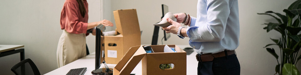 Young adult Caucasian man and young adult Caucasian woman cleaning office desks, packing personal belongings into cardboard boxes, organizing workspace during office move or cleanup