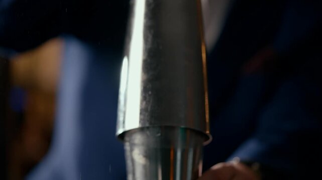 Bartender shakes a cocktail shaker with precision and focus. The metallic shaker gleams under warm lights, droplets of condensation form on its surface. The person wears a sharp suit