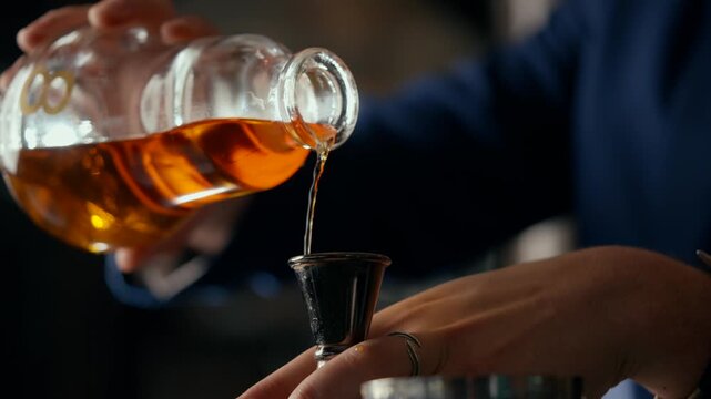 Hand pours amber liquid from a bottle into a small glass. The person's focus is precise and calm, suggesting care. The dim lighting highlights the rich color of the drink