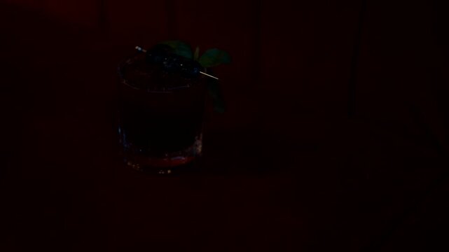 Cocktail in glass rests on velvet sofa. Red drink with berries and mint garnish. Warm lighting enhances cozy, inviting mood. Perfect for bar, lounge, or advertising use