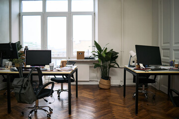 Fototapeta premium Empty modern office showing clean desks with computer monitors, organized workspace and polished wooden floor, large windows providing natural light, indoor plants adding greenery