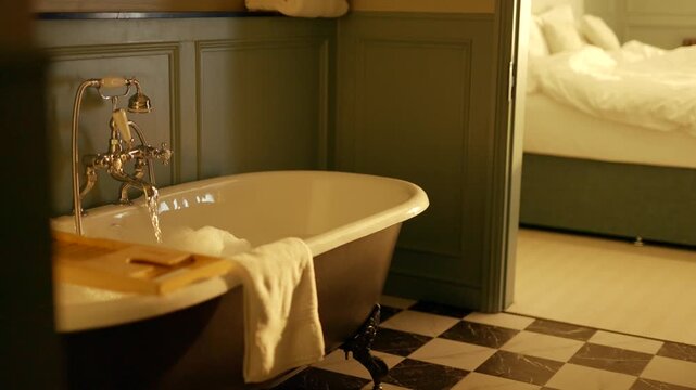 Vintage clawfoot bathtub with checkered floor. Soft light glows warmly in quiet, elegant bathroom. Towel draped over tub edge, soap on wooden tray. Bedroom visible in background with soft bed