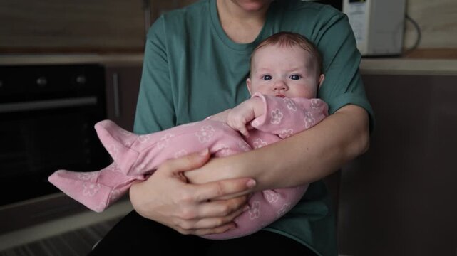 A mother holds her infant in her arms in a kitchen setting. The baby in a pink romper looks at the camera