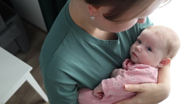 A woman in a green t-shirt holds a conscious baby in her arms. The infant in pink floral clothes looks towards the camera. Close-up view in natural light
