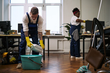 Young adult Caucasian man pouring cleaning solution into bucket while young adult Black woman wiping computer monitor in modern office workspace during cleaning routine © DragonImages
