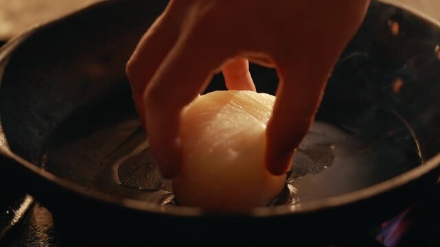 Hand gently places a scallop into hot oil in a cast iron pan. The scallop sizzles as it begins to cook. Smoke rises, highlighting the searing action. This close-up captures the texture