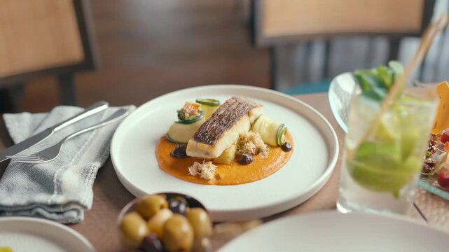 Beautifully plated fish dish with orange sauce and vegetables. Olives, garnishes, and a refreshing drink complete the table. Warm tones suggest a cozy, upscale dining experience