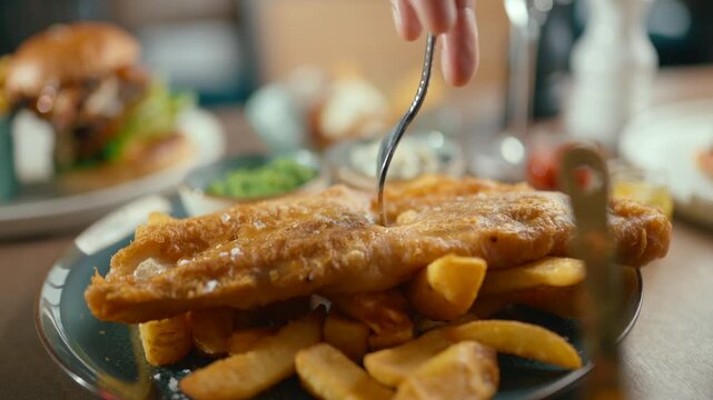 Golden-brown fish fillet with crispy batter sits atop golden fries. Hand uses a fork to lift the fish. Background shows blurred burger and condiments. Footage captures classic British fish