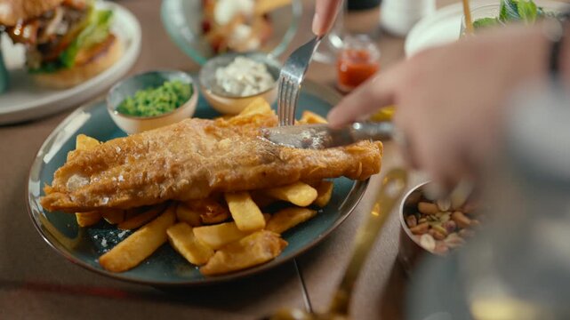 Person cuts golden fried fish and chips with fork and knife. Plate features crispy fries and fresh condiments. Scene shows cozy, casual dining with warm lighting. Ideal for food blogs