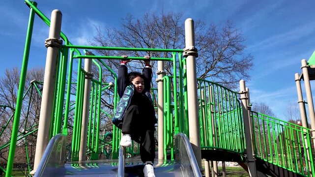 A joyful 4-year-old child swings from monkey bars above a slide at a sunny neighborhood park.