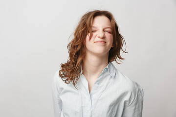 Young woman with curly hair wearing white shirt makes funny face with closed eyes and scrunched...