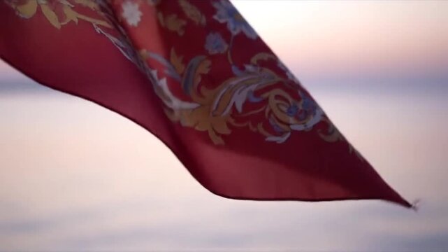 A red patterned scarf flutters in the wind against a soft, blurred sunset or sunrise background over a body of water, creating a calm and flowing scene.