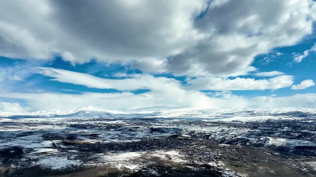 Vast snowy valley under dramatic cloudy sky. Mountains stretch far into distance. Snow blankets rugged terrain, forests below. No people visible in scene. Ideal for winter travel, adventure