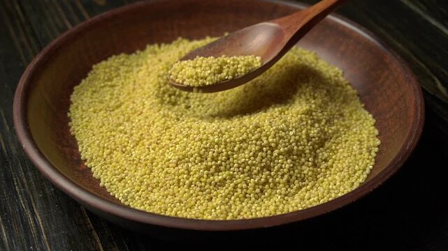 Yellow millet grains in a wooden bowl with a wooden spoon stirring the grains, showcasing the texture and color of the food product