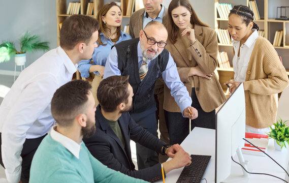Mentor teacher guides students during team training. A group gathers at a computer in a bright office, focused on learning, teamwork and skills. Concept of workplace learning and teamwork.