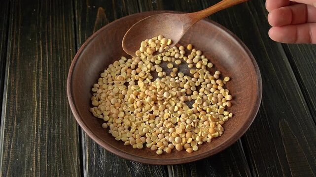 A hand in motion with a wooden spoon pouring yellow split peas into a wooden bowl as the grains scatter across a rustic wooden surface, highlighting the texture and color of the peas