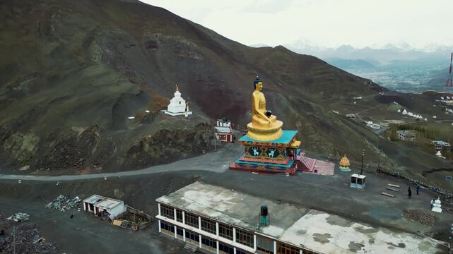 A cinematic drone orbit captures the towering golden seated Buddha statue as it overlooks the rooftops of Stok, with the dramatic snow-covered peaks of the Ladakh Range stretching across the horizon.