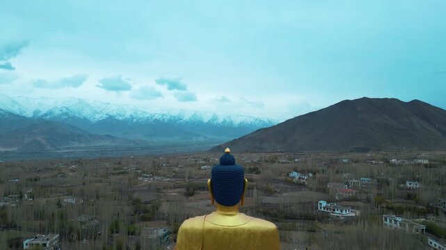 A cinematic drone orbit captures the towering golden seated Buddha statue as it overlooks the rooftops of Stok, with the dramatic snow-covered peaks of the Ladakh Range stretching across the horizon.