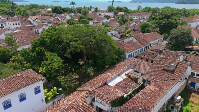 Paraty historic center showing blue and white colonial style facades and clay roofs, with Bahia Carioca bay and ocean visible in the background. Slow aerial glide in 4K.
