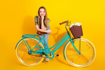 Young stylish woman with teal bicycle and basket of flowers posing against a bright yellow...
