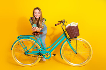 Young chic woman with turquoise bicycle and basket of flowers smiles against a bright yellow...
