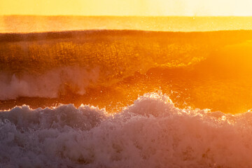 Fototapeta premium Waves crash onto the shore during sunrise, creating a dynamic scene with sea spray and mist. The golden light of the rising sun illuminates the ocean, giving the spray a vibrant glow.