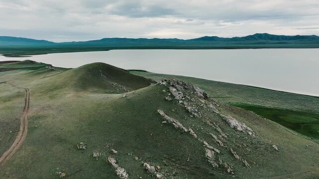 Rocky green hills near a large body of water under a cloudy sky, natural landscape with distant mountains and calm water.