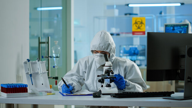 Biologist in ppe suit working on microscope and writing information on clipboard in equipped laboratory. Chemist examining vaccine evolution using high tech researching diagnosis against covid19 virus