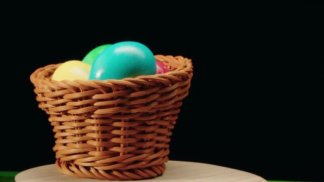 Rotation around axis of wicker basket with painted colorful easter shiny eggs on wooden surface against black background. Closeup studio shot emphasizing woven texture. Organic natural food