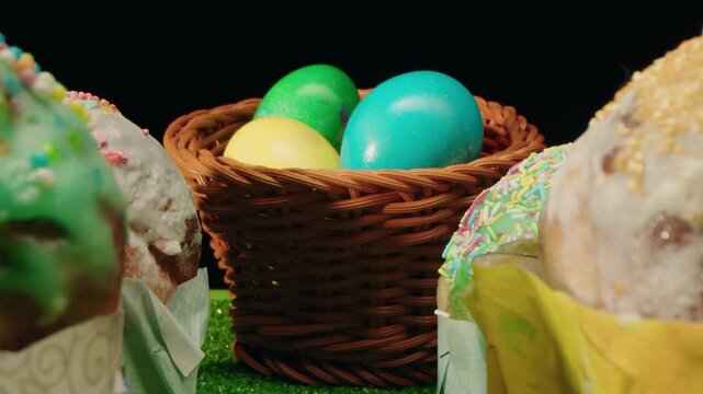 Closeup of easter turquoise and yellow eggs in wicker basket and glazed cakes with colorful sprinkles on soft icing. Spring composition on green grass and dark background. Macro of festive symbols