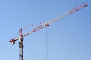 A large red and white industrial tower crane stands tall against a solid clear blue sky background at a construction site