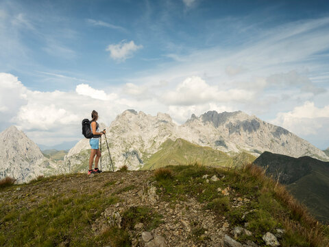 A woman with a backpack and hiking poles stands on a grassy mountain ridge, looking at the mountains in the distance, the Coglians gr under a blue sky. Monte Crostis,Carnia,Friuli Venezia Giulia,Italy