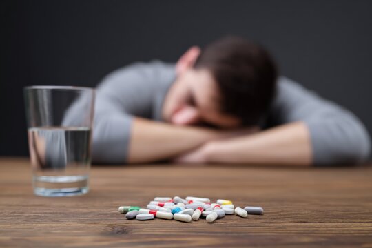 Young caucasian male adult leaning on table with glass of water and scattered pills in foreground suggesting fatigue and medication misuse