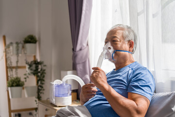 Asian senior man retirement using nebulizer mask for breathing treatment at home. Elderly patient...