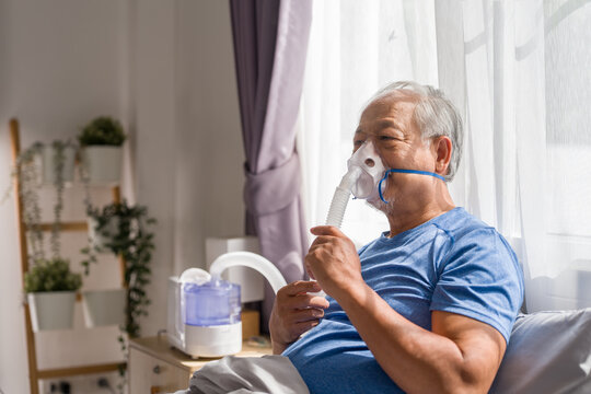 Asian senior man retirement using nebulizer mask for breathing treatment at home. Elderly patient with respiratory problem sitting on bed oxygen therapy pulmonary rehabilitation healthcare concept