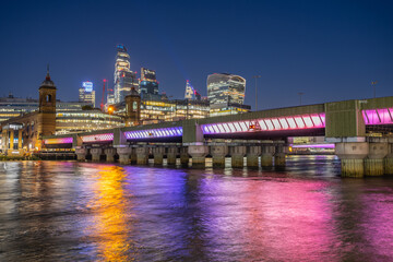 Cannon Street Railway Bridge stands over the water at night. The bridge is lit with colorful lights. The skyline of London features tall buildings in the background. © pyty