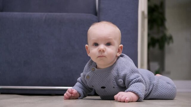 Adorable infant boy with blue eyes wearing a grey jumpsuit learning to crawl on the floor of a cozy living room. Cute toddler exploring the world and developing motor skills at home