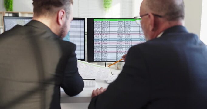 Accountants conduct analysis on dual monitors. Two colleagues review spreadsheets and discuss metrics, checking budgets and cashflow for accurate reporting. Teamwork in finance clearly shown.