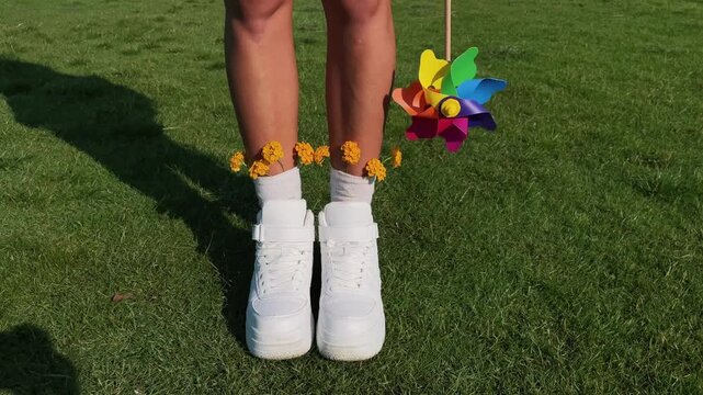 Horizontal video of female legs in white sneakers and socks in a flower field while a colorful rainbow pinwheel spins in the wind. Sunny day, playful mood. Concept of spring, summer, childhood, freedo