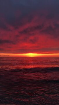 Dramatic red sunset sky over the calm sea. The horizon glows with deep crimson and orange tones as the sun disappears, reflecting on the water surface. 