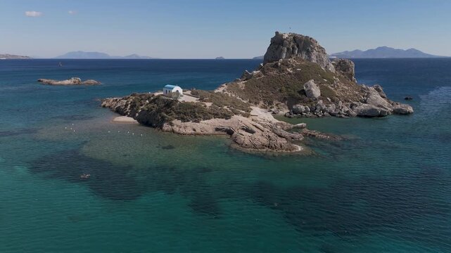Drone aerial view of white and blue small church in an island in front of Agios Stefanos Beach in Kos Greece