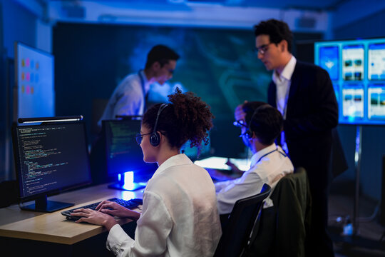 Professional male software engineer pointing at computer screen to help female programmer. IT development team working on coding data analysis in dark cyber security workplace.
