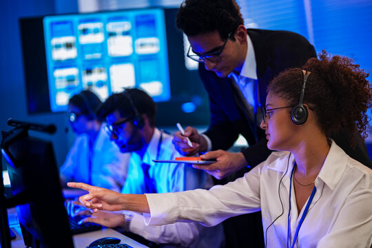 Diverse IT helpdesk team wearing headsets monitoring network operations. Tech professionals collaborating on digital software support in dark 24/7 cyber workspace.